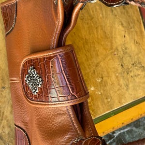 May include: A brown leather handbag with a crocodile-textured flap and accents. The bag features a braided handle and silver-toned hardware. The design includes decorative stitching and a silver emblem on the front. The bag's dimensions are not visible.