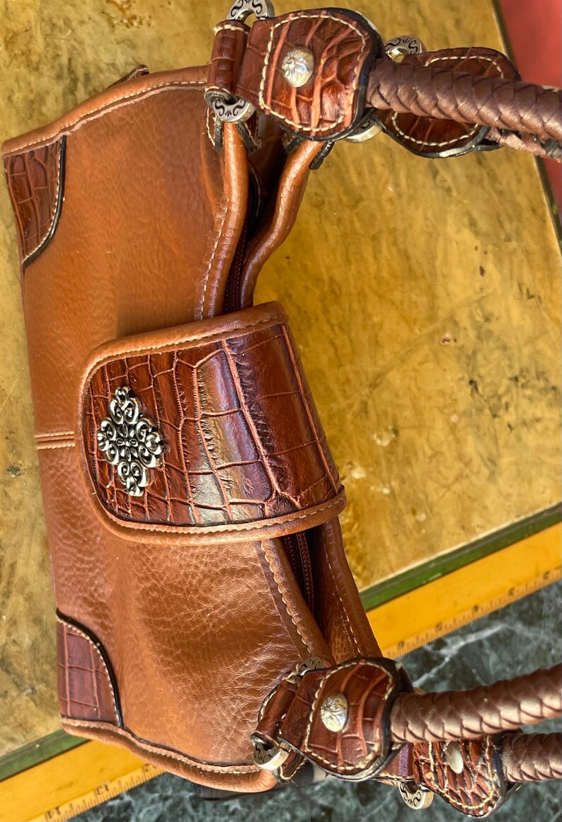May include: A brown leather handbag with a crocodile-textured flap and accents. The bag features a braided handle and silver-toned hardware. The design includes decorative stitching and a silver emblem on the front. The bag's dimensions are not visible.