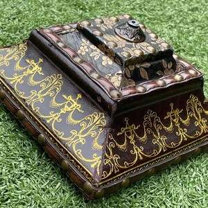 May include: A decorative, antique-style box with a dark brown leather exterior. The box features gold-colored ornate designs and is adorned with small, brass-colored studs. The top section is black with floral accents and a metal fixture.