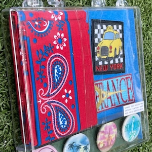 May include: A clear plastic travel-themed case containing a red paisley bandana, patches with a yellow taxi and the text "NEW YORK", and "FRANCE", plus four round pins with travel-related images and text. The case has a clear handle.