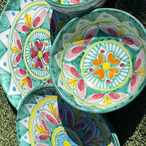 May include: A set of hand-painted ceramic bowls and plates with a vibrant floral design. The bowls and plates feature a teal green background with yellow, pink, and orange floral patterns. The set includes various sizes of bowls and plates.