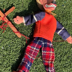 May include: A vintage puppet with a straw hat, a painted face, and a red and orange outfit. The puppet's pants are red, white, and blue plaid. The puppet is lying on green grass, with its wooden control cross visible.