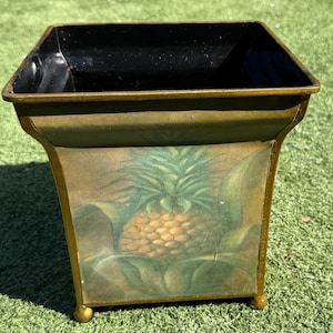 May include: A square metal planter with a painted pineapple design. The planter has a black interior, gold trim, and small gold feet. The pineapple is depicted with green leaves and a yellow-brown fruit.