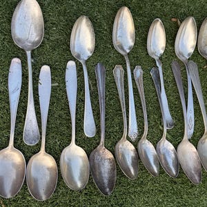 May include: An assortment of antique silver spoons of various sizes and designs, arranged on a green surface. The spoons have different handle styles and bowl shapes, showing signs of age and use. A yellow ruler is visible.