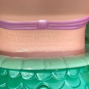 Pode incluir: Close-up do pesco&ccedil;o e ombros de uma boneca. A boneca tem um pesco&ccedil;o rosa claro com uma faixa roxa. A saia verde da boneca &eacute; vis&iacute;vel na parte inferior. O texto na boneca diz "TOLLYTOTS LIMITED MADE IN CHINA FABRIQUE EN CHINE Information at www.tollytots.com Part No: 01721-1-D01-12-02."
