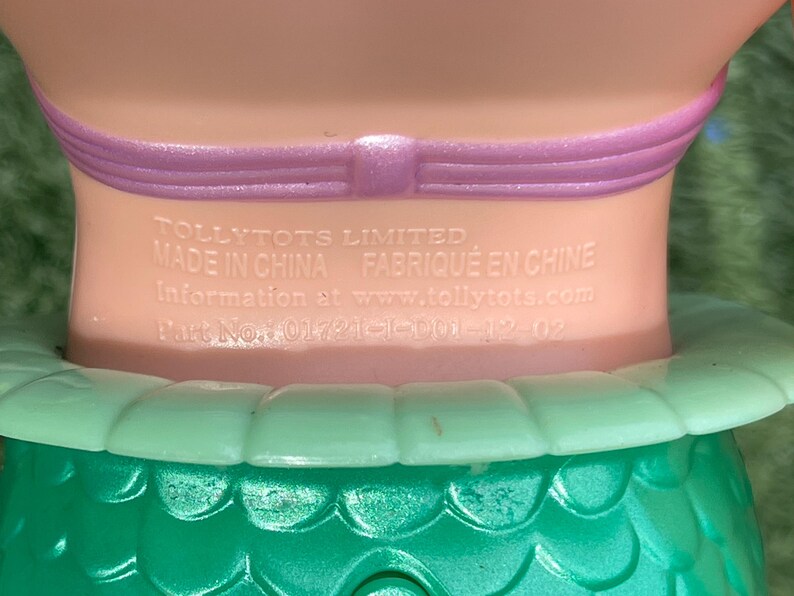 Pode incluir: Close-up do pesco&ccedil;o e ombros de uma boneca. A boneca tem um pesco&ccedil;o rosa claro com uma faixa roxa. A saia verde da boneca &eacute; vis&iacute;vel na parte inferior. O texto na boneca diz "TOLLYTOTS LIMITED MADE IN CHINA FABRIQUE EN CHINE Information at www.tollytots.com Part No: 01721-1-D01-12-02."