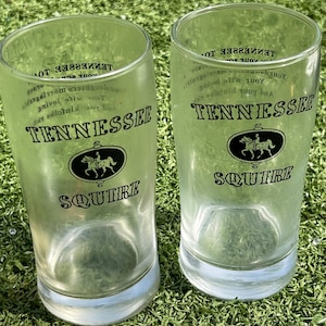May include: Two clear glass tumblers with the text "TENNESSEE SQUIRE" in black. Each glass features a black oval emblem with a horse and rider. The glasses are on a green surface.