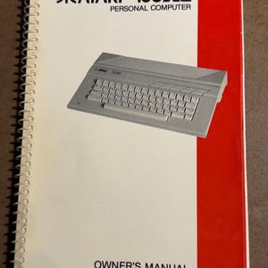 May include: An Atari 130XE Personal Computer owner's manual. The cover is white with the Atari logo and model name in black. A red vertical stripe is on the right side. The manual features an illustration of the computer.