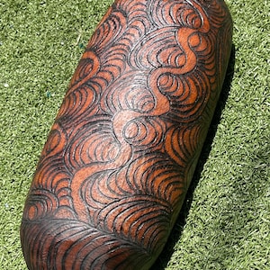 May include: A wooden, oblong sculpture with a dark brown finish and intricate, swirling patterns carved across its surface. The sculpture's design features a series of curved lines and shapes, creating a textured appearance. The piece is set against a backdrop of green.