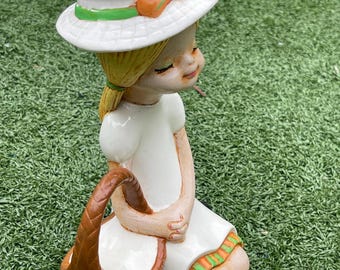 May include: A ceramic figurine of a girl kneeling on the grass, wearing a white dress and hat. The hat has an orange bow and green trim. She holds a brown basket. The figurine has blonde hair and a serene expression.