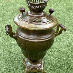 May include: Antique bronze samovar with a decorative top, spigot, and handles. The samovar has a round body and sits on a decorative base. The handles and top knobs are made of wood. The samovar is a traditional tea-making device.