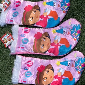 May include: Three pink Christmas stockings featuring Dora the Explorer. Each stocking has a white faux fur cuff and a cartoon image of Dora. The word "Dora" is printed vertically on each stocking, along with Christmas ornaments.
