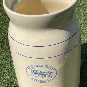 May include: Cream-colored ceramic water cooler with a wide opening and a spigot at the bottom. It features two blue stripes and a blue oval logo that reads "Mt. Rushmore Pottery Black Hills, S.D."