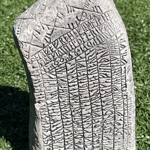 May include: A weathered, gray stone tablet with intricate carvings of ancient runes. The rectangular stone has a slightly curved shape and is placed on a green surface. The surface of the stone is covered in detailed, linear patterns.
