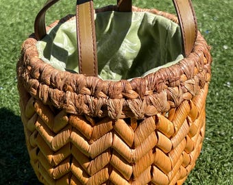 May include: A woven basket bag with a chevron pattern in shades of brown and tan. It features a green floral interior lining and brown leather handles. The bag is designed with a round shape.