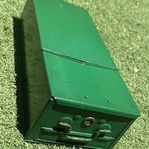 Op de afbeelding: Een rechthoekige groene metalen afsluitbare doos met een scharnierend deksel en een metalen handvat aan de voorzijde. De doos is voorzien van een centraal sleutelgatmechanisme en zichtbare metalen klinknagels langs de bovenrand.