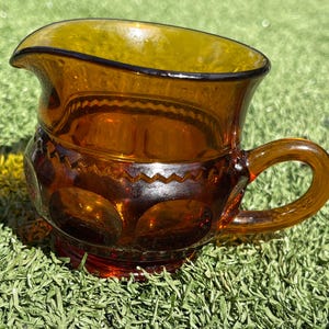 May include: Amber-colored glass creamer with a handle. The creamer has a decorative pattern around the middle and a flared top. The handle is rounded and attached to the side of the creamer. The creamer sits on a red base.