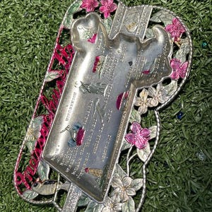May include: A silver-toned decorative tray with a rectangular central section and a floral border. The tray features a list of locations and is accented with pink and white flowers. The word "Winchendon" is visible on the tray.