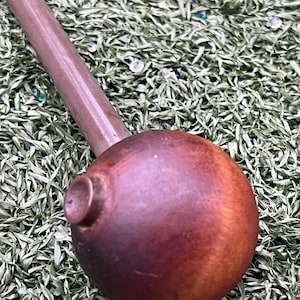 May include: A handcrafted, brown wooden pipe with a rounded bowl and a long, cylindrical stem. The pipe has a smooth, polished finish and a small, circular detail on the bowl. The pipe is set against a backdrop of green artificial grass.