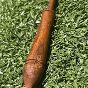 May include: A brown wooden drop spindle with a tapered, elongated body and a rounded base featuring carved horizontal grooves. The surface of the wood displays a natural grain pattern with a smooth finish and a small, faint carved emblem near the center.