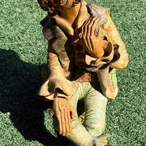May include: A carved figurine of a seated clown wearing a tall dark hat, a tan jacket with ruffled sleeves, and light green trousers. The figure has detailed facial features, curly hair, and brown shoes with green accents, rendered in a wood-like finish.