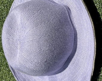 May include: A light purple sun hat with a wide brim. The hat is made of a woven material and has a rounded crown. The brim is slightly wavy and the hat is sitting on green grass.