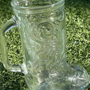 May include: Clear glass boot-shaped mug with a handle. The mug features embossed decorative scrollwork and a stitched design around the top and bottom. The mug is sitting on a bed of green grass.