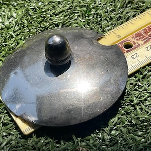 May include: A round, silver-colored metal lid with a small, dark knob on top. A yellow ruler with black markings is partially visible. The lid is approximately 3 inches in diameter.