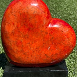 May include: A large, polished red heart-shaped sculpture. The stone heart has a glossy finish and sits on a small, black rectangular base. The sculpture is displayed on a bed of green grass.