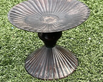 May include: A bronze-colored metal pedestal bowl with a fluted, fan-like design. The bowl has a wide, shallow dish supported by a dark, rounded stem and a flared base. The bowl is set on a green surface.