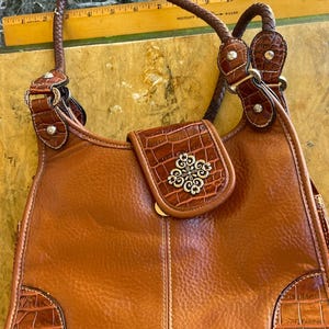 May include: A brown leather handbag with a braided handle and decorative silver-tone accents. The bag features a flap with a silver floral design and crocodile-textured leather details on the corners and flap.