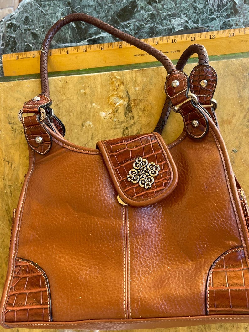 May include: A brown leather handbag with a braided handle and decorative silver-tone accents. The bag features a flap with a silver floral design and crocodile-textured leather details on the corners and flap.