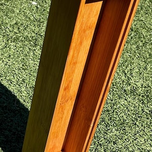May include: A rectangular bamboo box with an open top and a hollow interior. The box is a warm, natural wood tone and sits on a green, textured surface. The box is empty and has a simple, clean design.