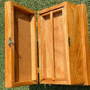 May include: Open wooden cabinet with a light brown finish. The cabinet has two doors, each with a decorative metal latch. The interior features a small shelf and vertical dividers. The cabinet is made of wood.