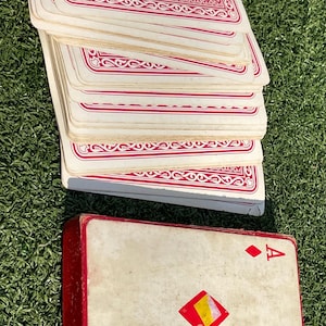 May include: A deck of playing cards with a red and white patterned design on the back. The Ace of Diamonds is visible on the card box. The cards are stacked on a green surface.