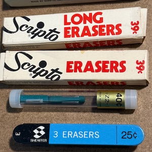 May include: A collection of vintage erasers and eraser packaging. Includes boxes of Scripto Long Erasers, a tube of green erasers, and a blue Sheaffer 3 Erasers package. The items are on a brown surface.