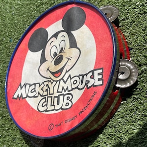 May include: Vintage Mickey Mouse Club tambourine. The red and white drum features Mickey Mouse's face and the words "Mickey Mouse Club." The tambourine has a blue rim, metal jingles, and a red and green striped handle.