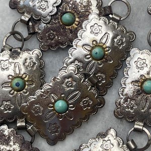 May include: Close-up of a silver-toned belt with turquoise-colored stone accents. The belt features a floral design and a scalloped edge. The metal has a distressed appearance.