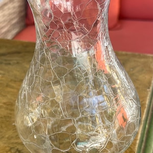 May include: Clear glass vase with a crackle finish. The vase has a bulbous base and a flared opening. The crackle pattern creates a unique texture. The vase is sitting on a wooden surface.