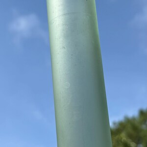 May include: A tall, cylindrical, frosted glass object. The object is a pale green color and has a slightly textured surface. The background is a clear blue sky with some green foliage visible at the bottom.