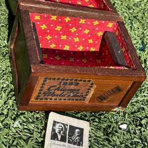 May include: A small wooden box with a hinged lid, lined with red fabric patterned with yellow flowers. The box has a label that reads "1893 Chicago World's Fair." A small paper with images is next to the box.