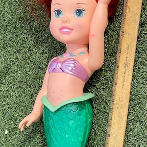 Pode incluir: Boneca de sereia com cabelo ruivo, olhos azuis e um top rosa. A boneca tem uma cauda verde e est&aacute; sobre relva verde. Uma r&eacute;gua de madeira est&aacute; ao lado da boneca.