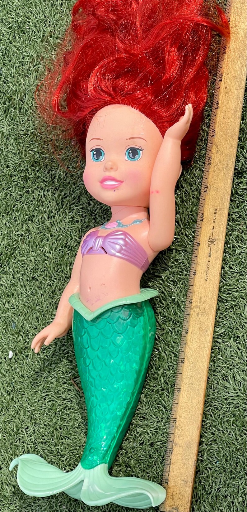 Pode incluir: Boneca de sereia com cabelo ruivo, olhos azuis e um top rosa. A boneca tem uma cauda verde e est&aacute; sobre relva verde. Uma r&eacute;gua de madeira est&aacute; ao lado da boneca.