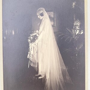 May include: Vintage black and white photograph of a bride in a long, flowing wedding dress with a veil. She holds a bouquet of flowers and stands in a room with a painting on the wall. The photograph is in a cream-colored frame.