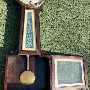 May include: Antique wooden wall clock with a brass-colored face and hands. The clock has a decorative blue and gold panel. The pendulum is visible, and the clock's door is open, revealing a framed picture. A vintage timepiece.