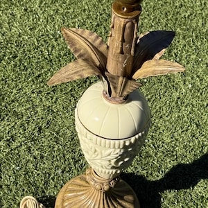 May include: A vintage table lamp with a cream-colored ceramic base and a gold-toned metal top. The base features a detailed leaf pattern and a round, scalloped foot. The lamp has a standard US plug and cord.