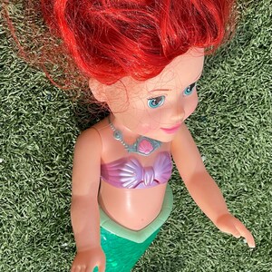 Pode incluir: Boneca de sereia com cabelo vermelho brilhante, olhos azuis e top rosa. A sereia tem uma cauda verde e um colar. A boneca est&aacute; sobre um leito de relva verde.