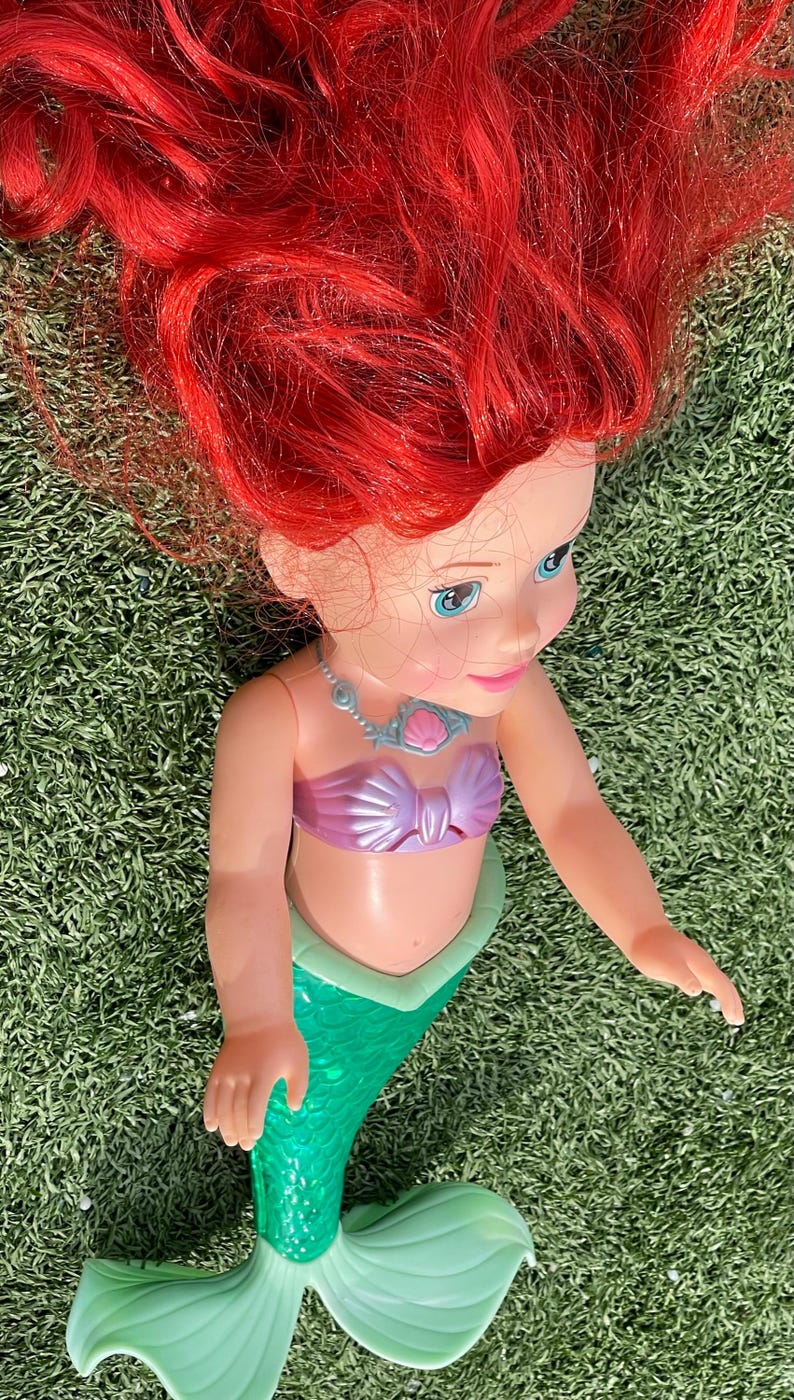 Pode incluir: Boneca de sereia com cabelo vermelho brilhante, olhos azuis e top rosa. A sereia tem uma cauda verde e um colar. A boneca est&aacute; sobre um leito de relva verde.