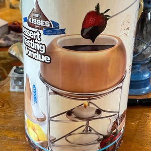May include: Hershey's Kisses dessert fondue set. The ceramic pot sits on a metal stand with a candle warmer. A strawberry dipped in chocolate is shown. The packaging features the Hershey's Kisses logo and the words "Milk Chocolate."