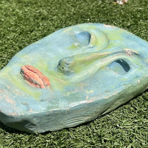 May include: A hand-painted, abstract sculpture in shades of blue, green, and orange. The piece has a textured surface with organic shapes and three openings. The sculpture is placed on a bed of green artificial turf.